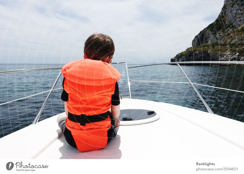 Rear view of a young boy sitting on the deck of a motor boat and wearing an orange life jacket Boy child rear view motorboat buoyancy aid sea at sea facing away