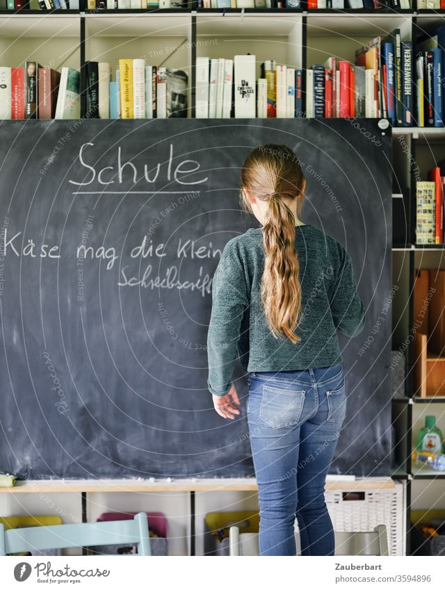 Homeschooling I - Girl writes with chalk on a blackboard a practice set for cursive writing, in the background bookcase School girl pupil schuler