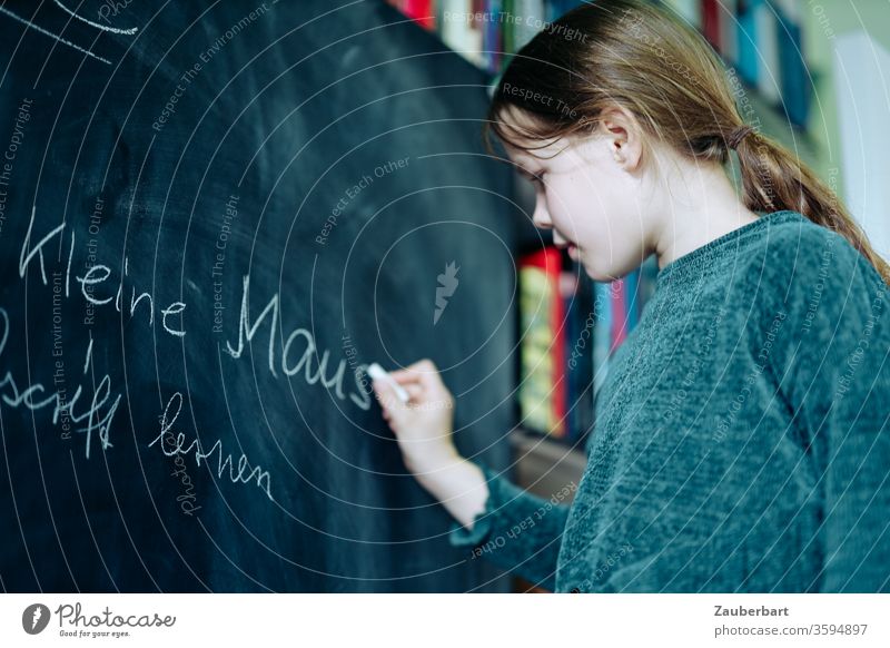 Homeschooling II - Girl writes with chalk on a blackboard a practice set for cursive writing, in the background bookcase School girl pupil schuler