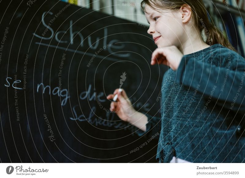 Homeschooling VII - Girl plays school and writes with chalk on a blackboard a practice set for cursive writing, in the background bookcase School girl pupil