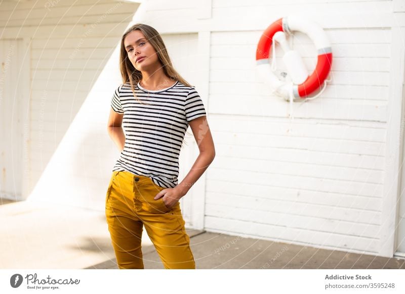 Stylish woman on quay near building with lifebuoy lifesaver style trendy stripe port calm female wall wooden stand young confident summer relax fashion content