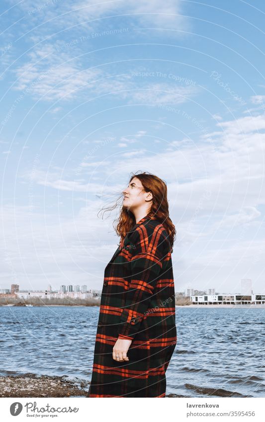 Lifestyle portrait of the young adult red-haired woman in checked coat on the bank of a river or sea, selective focus water bright white travel pretty beach
