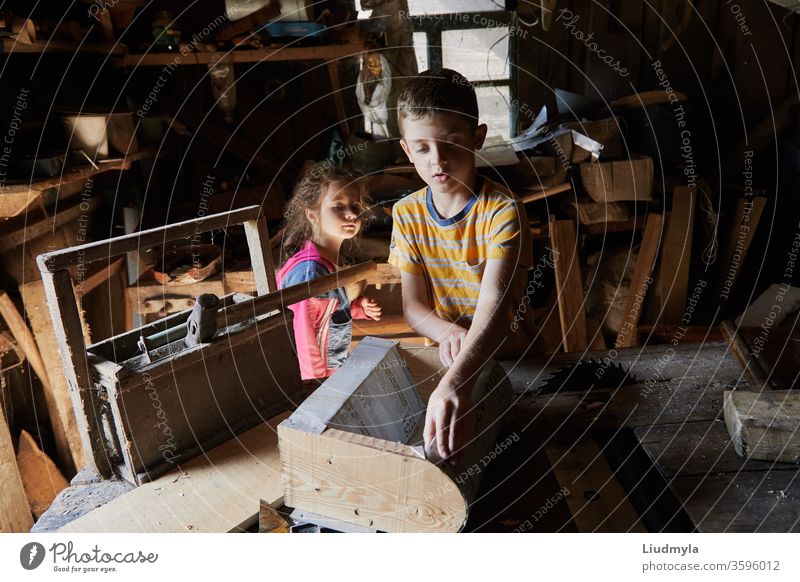 Young boy is making a wooden  bird house in the carpenter workshop. Curious little sister is trying to see what he is doing behind his back curious handyman