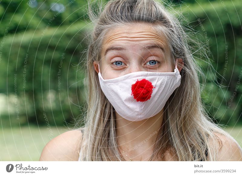 Young pretty woman looks anxiously into the camera with wrinkled forehead and mask. Red Nose Day with funny, funny clown mask, with red bobble, pompom to protect against corona. Blonde, young, long-haired woman with cool face mask outside in the nature.