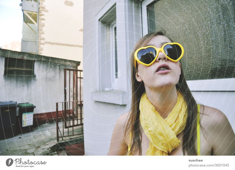 teenager with yellow heart sunglasses and yellow scarf in yellow bikini stands in a somewhat run-down backyard and looks up girl Young woman