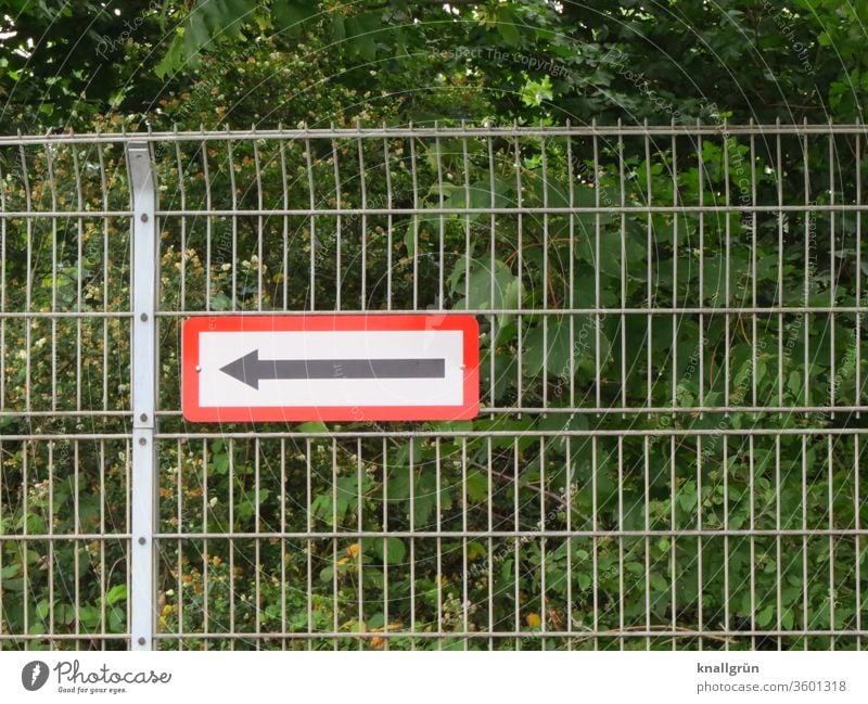 Grid fence with sign with arrow pointing to the left. In the background green bushes and trees. Arrow Fence Signs and labeling Signage Direction Orientation
