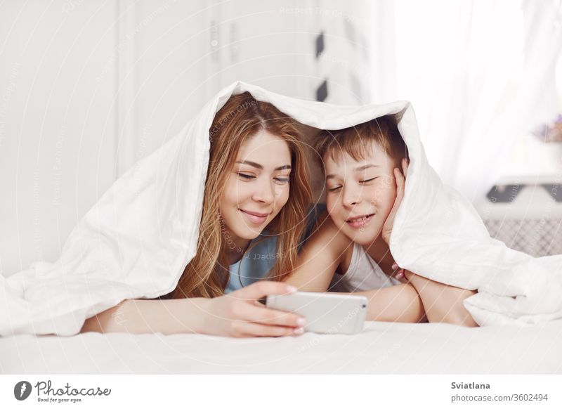 A beautiful young girl and her cheerful younger brother look at photos on their phone while lying on the bed with a smile. Brother and sister watch videos on their phone.
