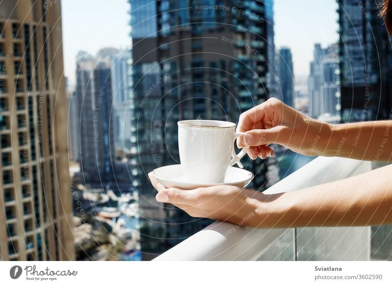 A girl holds a Cup of coffee against the background of modern buildings. A woman drinks morning coffee on the balcony during self-isolation city luxury view