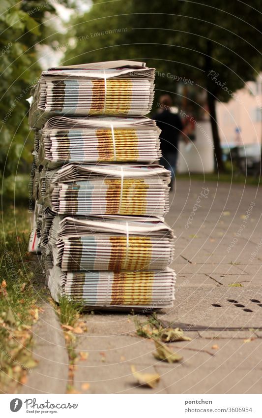 Stacks of weekly newspapers in bundles wait on the sidewalk for the delivery person. In the background a person moves away. sideline Pensioner job student job
