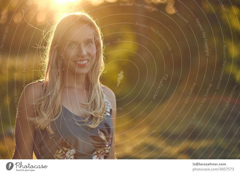 Smiling attractive blond woman backlit at sunset by the warm glow of the sun through leafy green trees in a blurred rural background with copy space sunrise