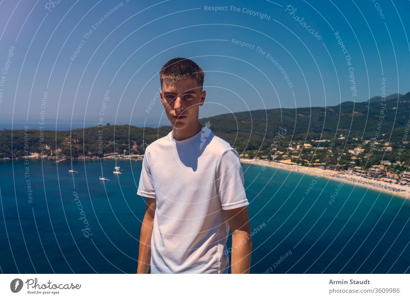 portrait of a self-confident young man with the beach of the city of parga in the background beautiful blue boy casual caucasian cloudless journey lifestyle