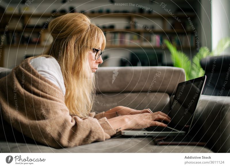 Pensive woman working on laptop at home browsing freelance project using telework lying sofa remote female serious eyeglasses gadget internet device computer