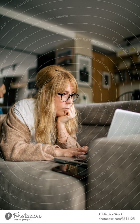 Pensive woman working on laptop at home browsing freelance project using telework lying sofa remote female serious eyeglasses gadget internet device computer