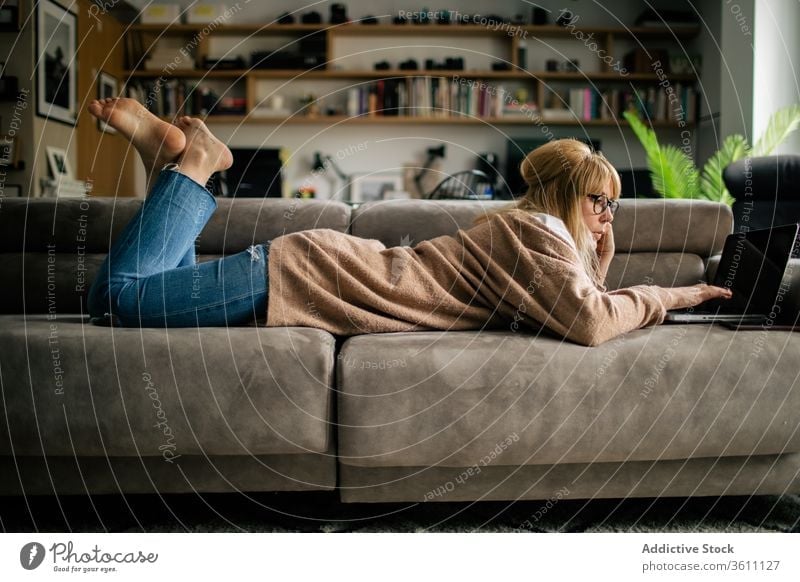 Pensive woman working on laptop at home browsing freelance project using telework lying sofa remote female serious eyeglasses gadget internet device computer