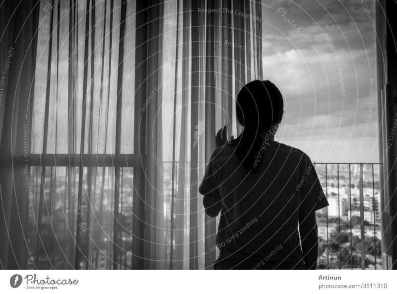 Sad adult Asian woman looking out of window and thinking. Stressed and depressed young woman. Despair women with long hair and T-shirts standing by the window of home or hospital. Lonely and anxiety.