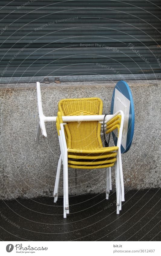 Yellow, gaudy, trendy plastic bistro chairs are stacked on top of each other, in front of a grey, dreary house wall, with closed shutters. End of work, end of season in a street café with furniture, chairs and blue, round bistro table stacked on top of each other. To