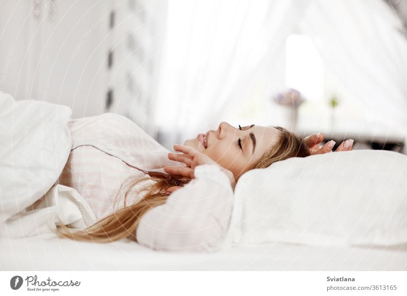 A young smiling beautiful girl is lying in bed in the bedroom. The girl stretches in the morning after waking up. Side view, space for text eyes closed indoors