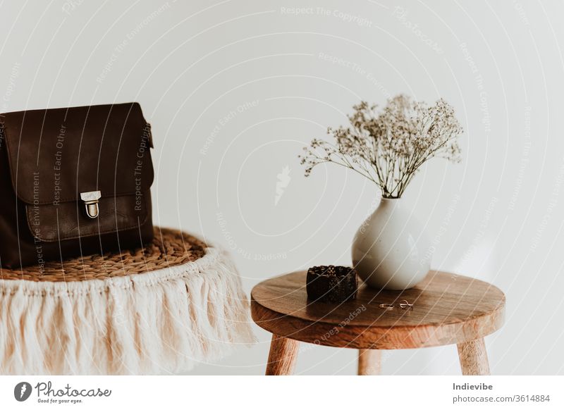 Accessory composition detail for women and girls. White flower in a small ceramic vase with copper bracelet and golden rings and leather bag on a wooden side table at a white wall in a bright room.