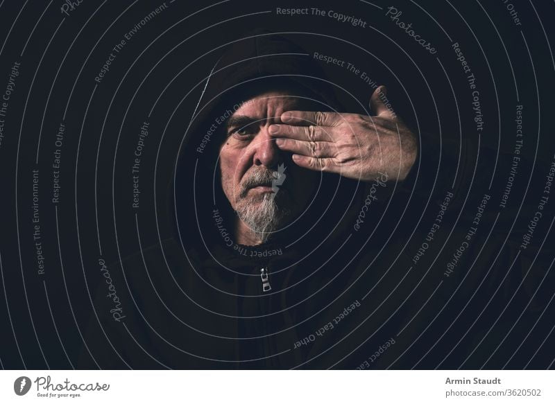 portrait of a man wearing a hoodie, holding one hand in front of an eye blind see cover finger beard gray glass black dark serious danger intensive caucasian