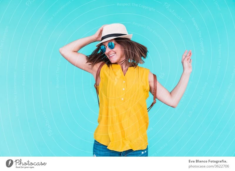 portrait of young woman dancing and jumping outdoors over turquoise background. Summer time mobile phone summer sunglasses yellow hat backpack city urban