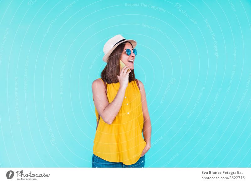 young woman using mobile phone outdoors over turquoise background. Summer time summer sunglasses yellow hat backpack city urban lifestyle happy smiling girls