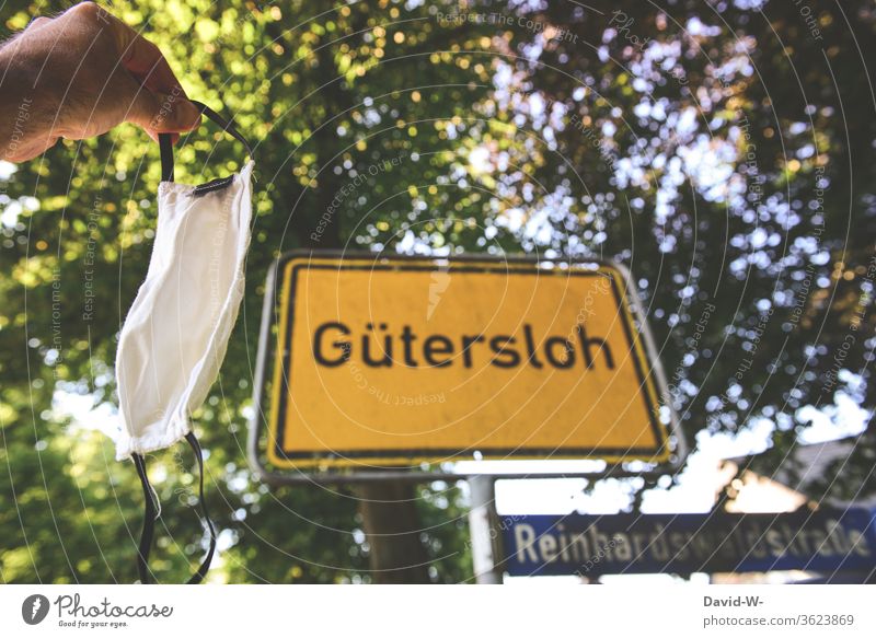 Man holds a breathing mask in front of the place-name sign in Gütersloh - Corona / covid-19 place-name sign - Gütersloh Rheda-Wiedenbrück Oelde cordon
