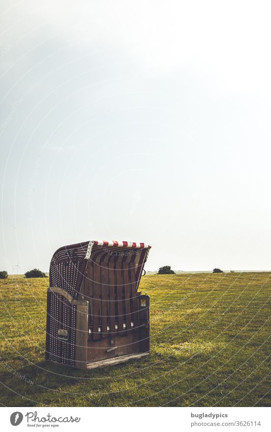 Red and white striped wicker beach chair on the dyke/ a grass/ lawn. The basket is closed. In the background you can see a few flat bushes and a lot of bright/ pale sky. Muted colours. In the upper area there is space for text.