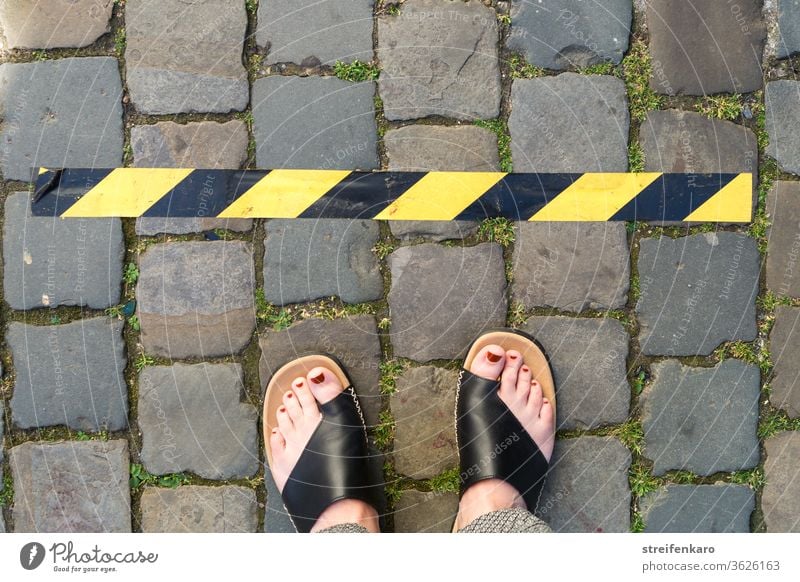 Keep your distance! - female feet stand in front of a black and yellow tape on cobblestone gap keep sb./sth. apart corona Corona virus Protection pandemic