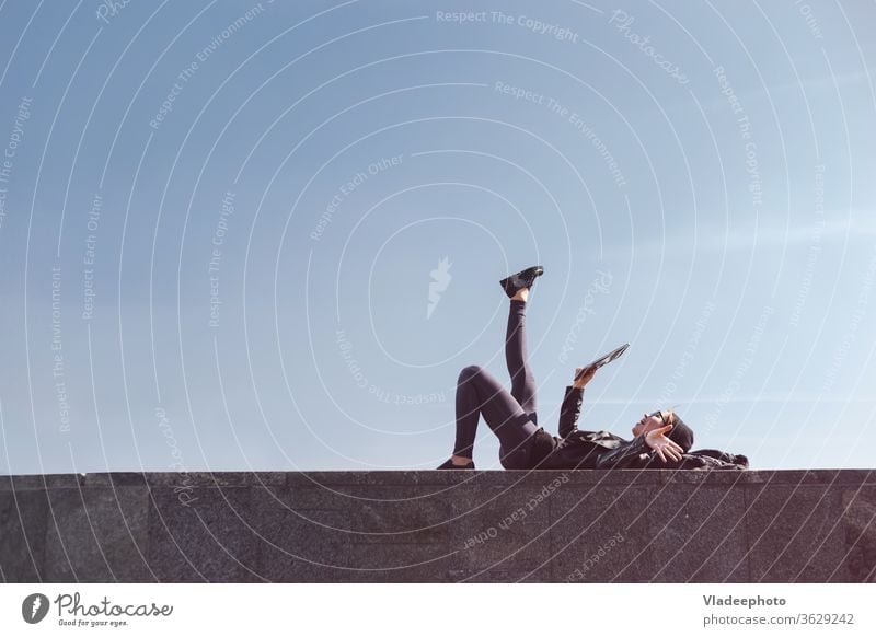 Side view of a teen using tablet on holidays lying outdoor with clear sky in background. Copyspace on top of frame. street music listening girl mobile summer