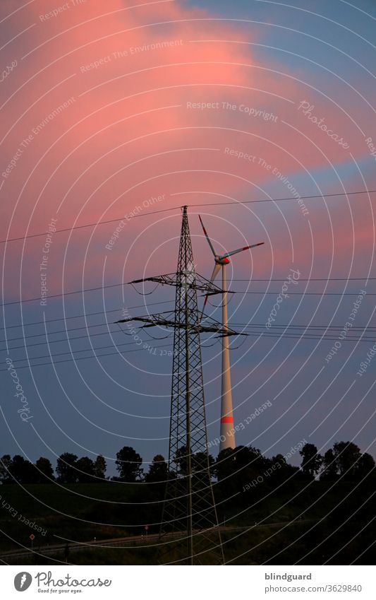 Energy mix. Windmill behind a mast of the overhead power line for the nationwide power supply, with forest in front of a red-blue sky in the evening.