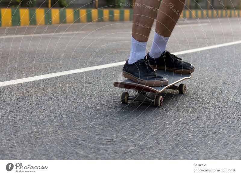 Close-up Shot of a Young Teenager's feet wearing Ripped Black Sneakers on his Skateboard cruising on an Empty Street while the White Line on the Road compliments his White Socks and also acts as a Leading Line in the Direction he is Moving.