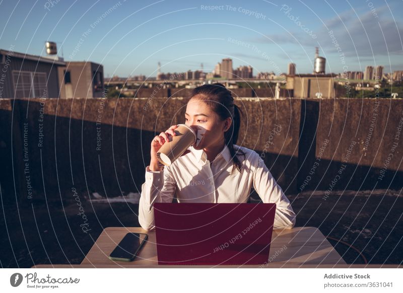 Young woman with laptop drinking coffee on rooftop work break busy pensive entrepreneur remote asian female young formal takeaway cup device gadget beverage