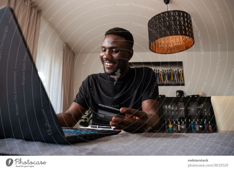 Young Black Man Smiling Online Shopping from Living Room credit card online shopping bank card man smiling happiness positive emotion purchase African ethnicity