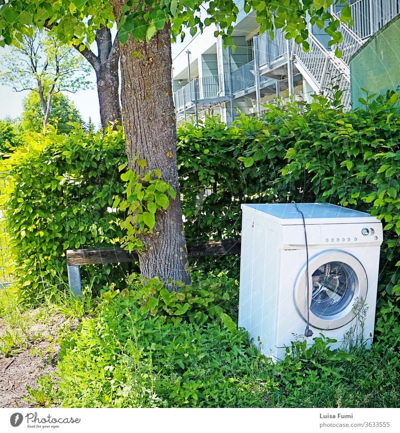 A washing machine dumped as garbage and abandoned on the sidewalk, sharply contrasting with the green environment discarded cityscape litter clean laundry tree