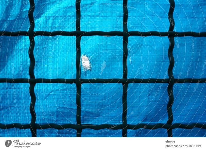 old lattice fence made of metal, backed with blue, coarse plastic foil as privacy protection, which has a crack as a peephole Metalware PVC tarpaulin Blue