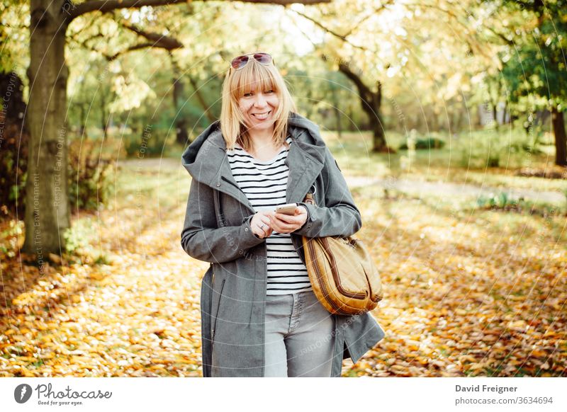 Happy blonde woman in a autumnal forest or park texting with her mobile phone. Communication, technology and outdoors concept. beautiful female happy person