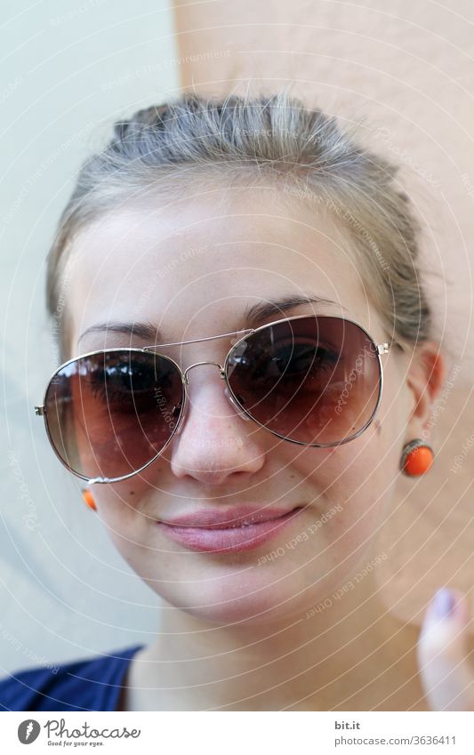 Teenager in front of a Mediterranean wall with orange earrings, ponytail and sunglasses. Teenager with blond hair smiles into the camera. Holiday photo of a young woman in summer, in front of a light-coloured facade, wall. Sun protection through crooked sunglasses.
