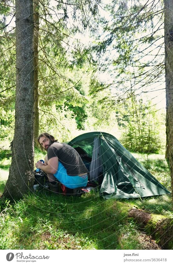 Man sits in the forest in front of a tent and looks back into the camera. Young man Tent Forest Morning Summer Sunlight Nature Adventure Vacation & Travel