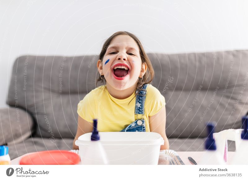 Expressive girl with dirty finger sitting on sofa at home grimace expressive mouth opened playful diy creative domestic child denim overall colorful amazed