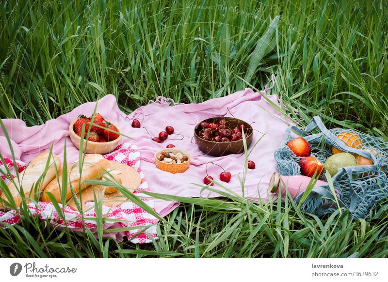 Zero waste summer picnic on the with cherries in the wooden coconut bowls, fresh bread and glass bottle of juice or smoothie on pink blanket, flatlay apples