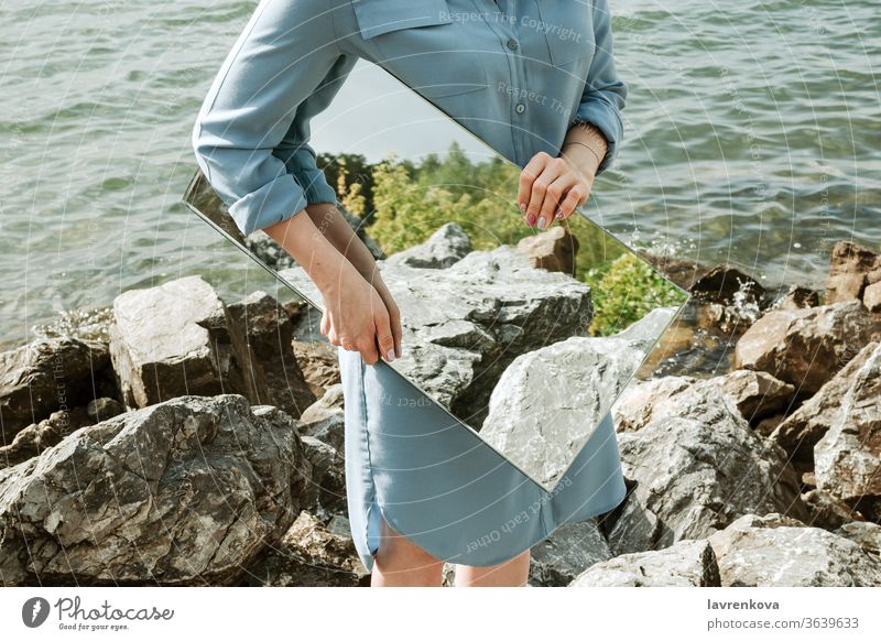 Closeup of woman standind on a shore in blue dress holding a mirror with trees and rocks reflection, selective focus adult clean concept environment faceless