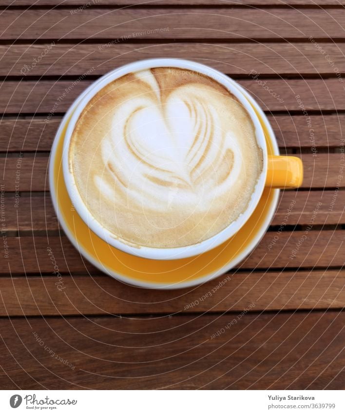 Yellow cup of coffe with milk on a dark background. Hot latte or Cappuccino decorated with foam with heart on a wooden table with copy space. Top view.