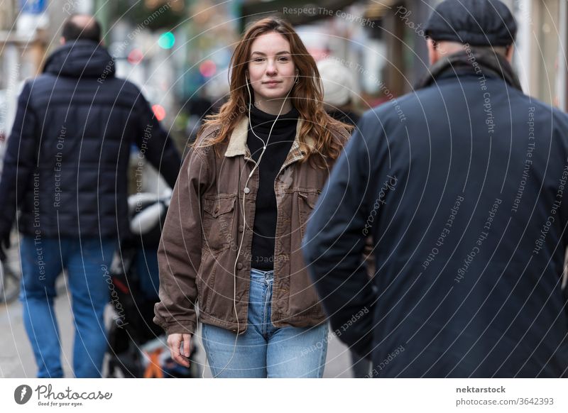 Young Woman Wearing Jeans and Brown Coat Walking Amongst Pedestrians caucasian ethnicity woman female earphones audio music listening sidewalk street
