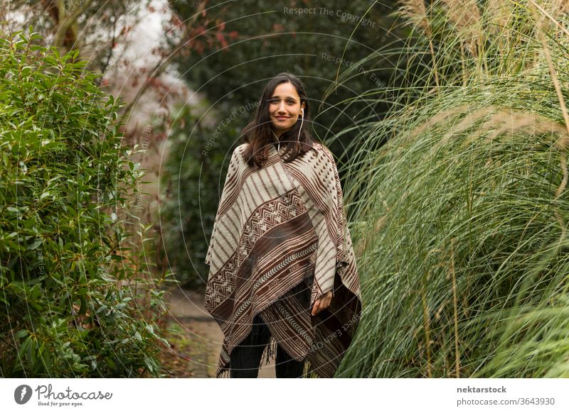 Young woman wearing poncho posing between tall grass in autumn. young woman Indian ethnicity middle eastern ethnicity earphones medium shot audiobook