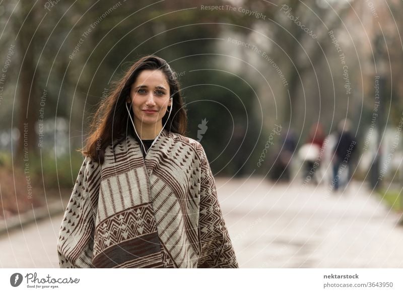 Young Indian woman wearing poncho and earphones smiling for the camera outdoors. Medium shot. young woman Indian ethnicity middle eastern ethnicity medium shot
