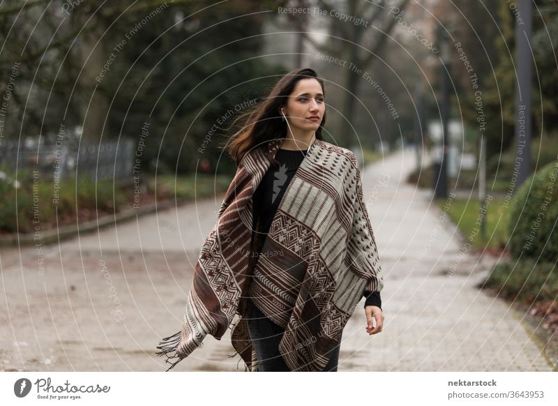 Young Indian woman wearing poncho walking outdoors with head turned looking away. young woman Indian ethnicity middle eastern ethnicity earphones medium shot