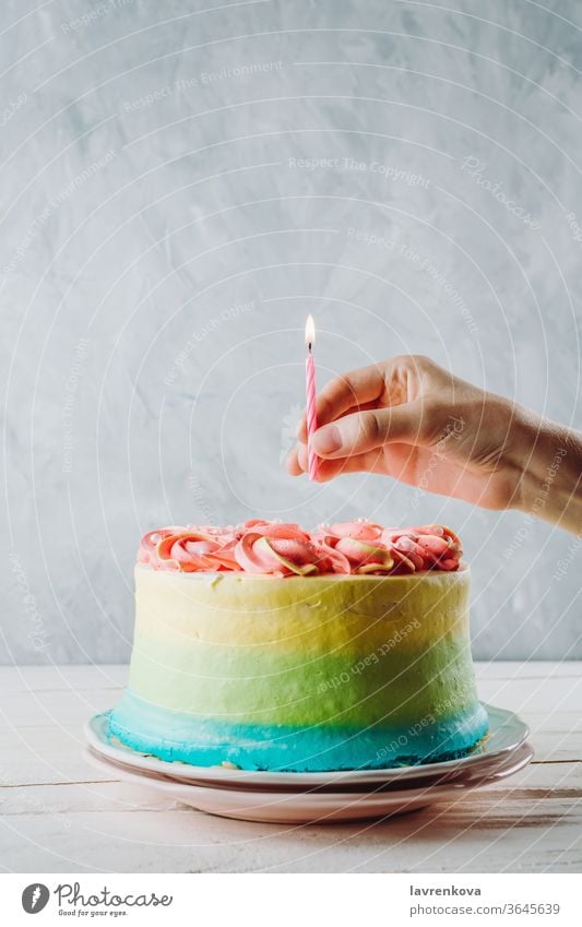Closeup of colorful vegan cake and woman's hand holding birthday candle above it, selective focus fingers hands female homemade fresh blue pink happy decoration