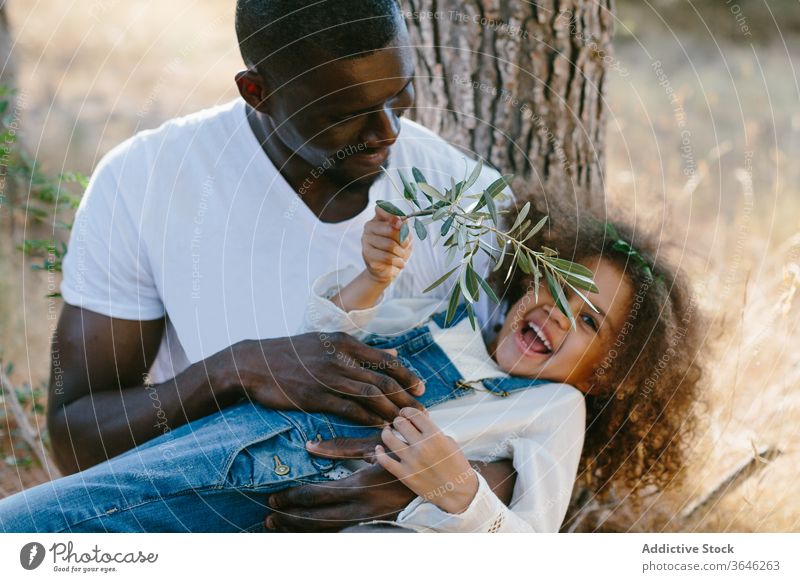 Happy African American father hugging daughter in nature tree mulatto black lap rest adorable summer positive forest smile casual happy countryside cute outfit