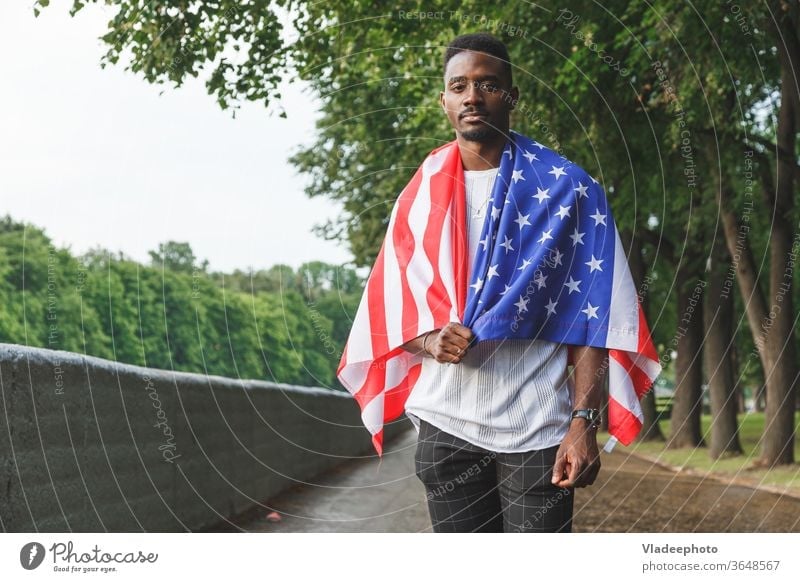 Handsome Afro American man with USA flag on his shoulders seriously looking at camera, standing outdoors. Day summer american male black african expat