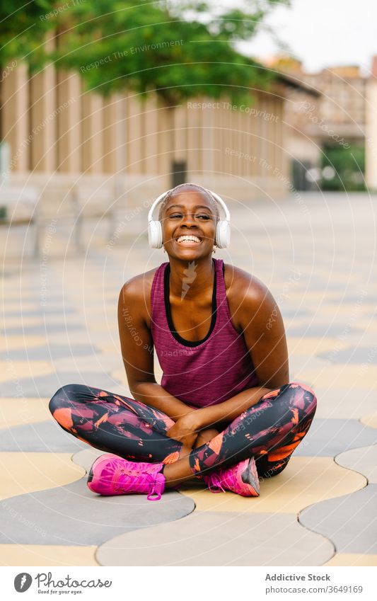 Happy black sportswoman in headset sitting on pavement after workout athlete sportswear embankment cheerful lean forward listen music healthy wellness vitality